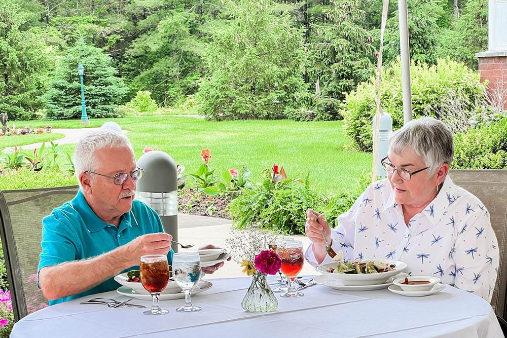 An elderly man and woman are dining outdoors at a table with plates of food, glasses of iced tea, and a vase of flowers, surrounded by greenery. This serene setting is part of their assisted living community's beautiful garden area.