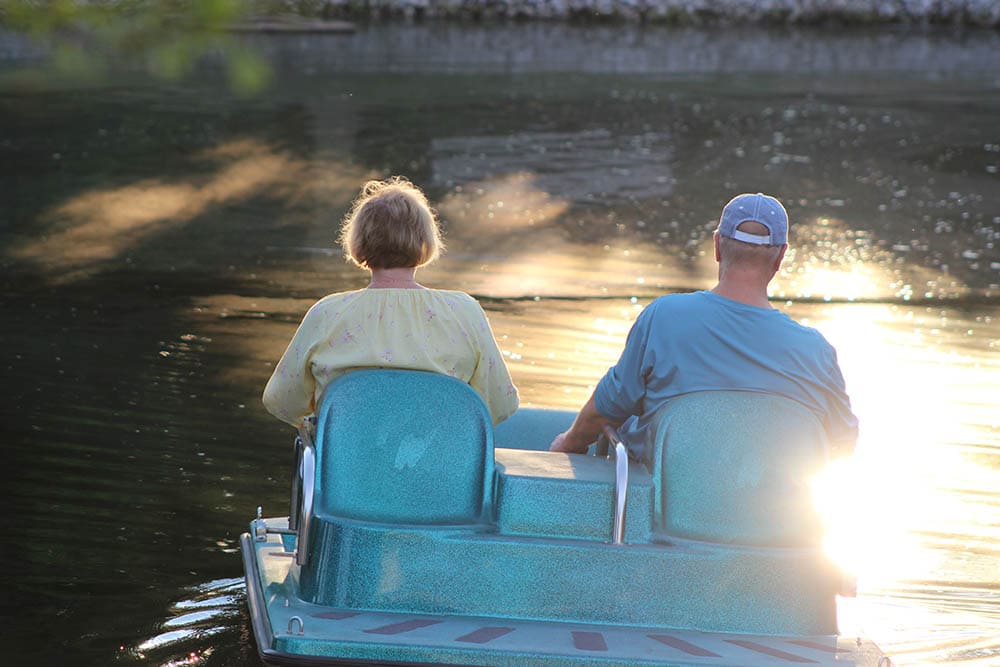 Two people are seated on a blue pedal boat on a calm body of water with sunlight reflecting off the surface. They are facing away from the camera, enjoying a serene day out from their senior living community.