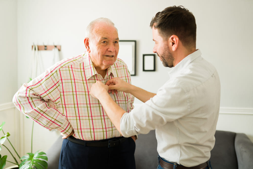 A younger man helps an older man button his plaid shirt in a living room. There is a gray sofa and some wall decorations in the background, showcasing the kind of compassionate care promoted by careers at United Church Homes.