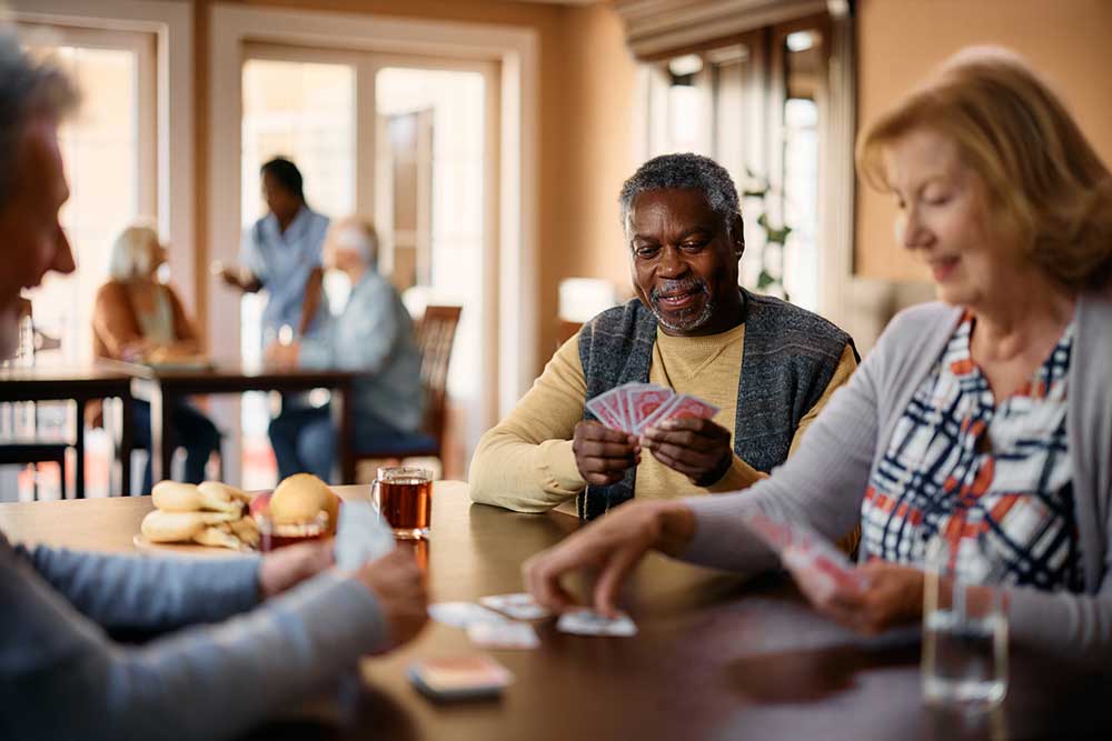 In a well-lit room, a group of older adults from the senior living community management team is seated at tables, engaging in card games and conversation. Food and drinks are visible on the table.