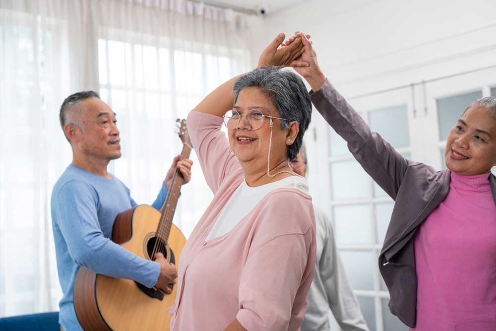 Three elderly people are enjoying a gathering; one man is playing a guitar, while two women dance together with smiles on their faces. The joy of the moment reflects their shared lifetime values, much like the thoughtful act of planned giving.