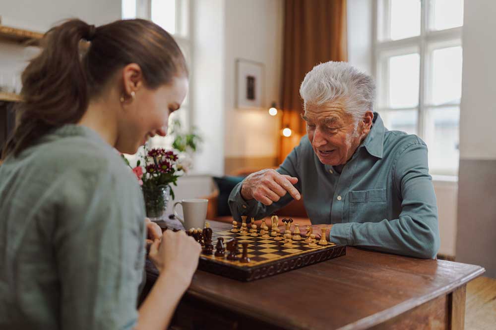 A young woman and an elderly man are playing chess at a wooden table in a well-lit room. The volunteer appears to be guiding or pointing out something on the chessboard.