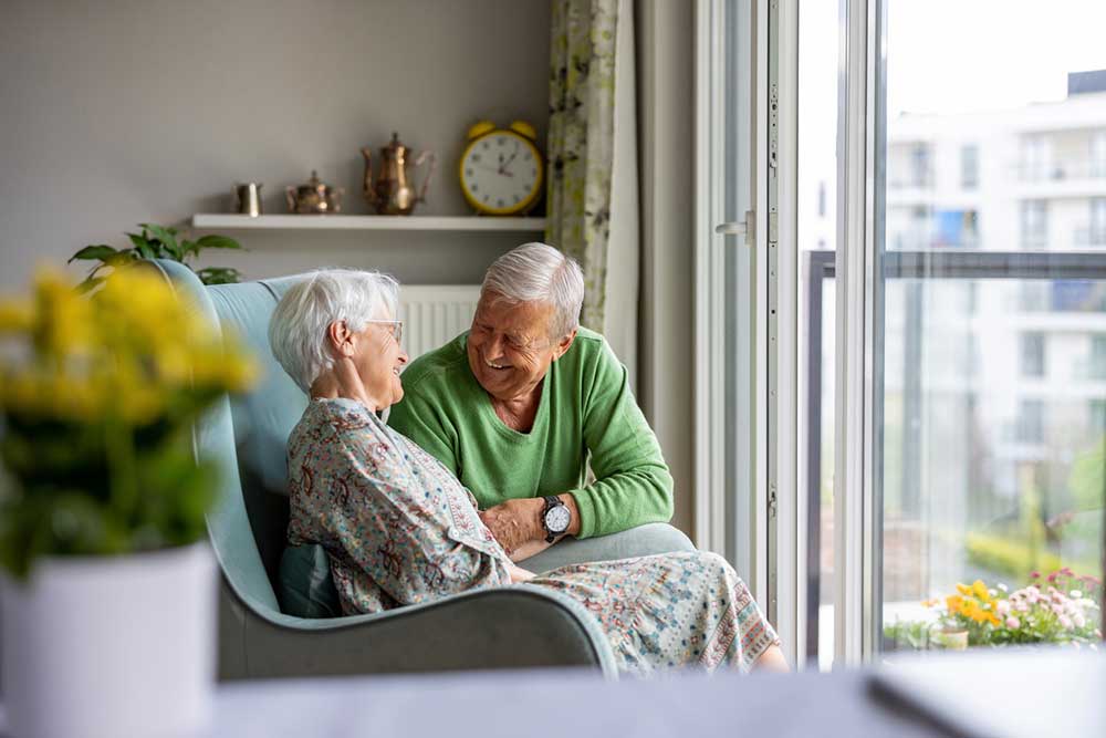 Two elderly people sitting together on a chair by a window, smiling and holding hands in a brightly lit room with a potted plant and clock visible in the background, enjoying their time with the reassurance of in-home senior health care.