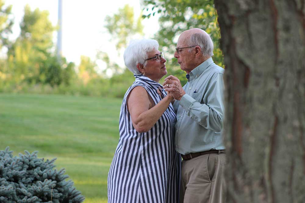 An elderly couple is dancing together outdoors on a grassy area, surrounded by greenery and trees. The woman wears a striped dress and the man wears a light-colored shirt and beige pants. The uch difference between their coordinated steps adds charm to the delightful scene.