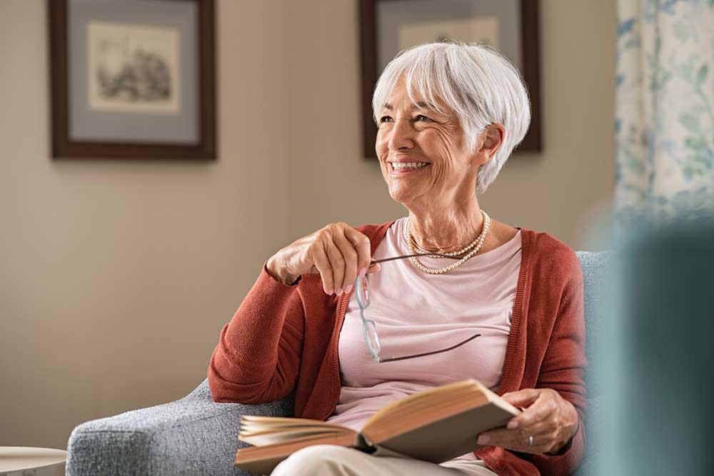 An elderly woman with short white hair sits on a chair, holding glasses and a book, and smiling. She wears a red cardigan and white shirt. Two framed pictures are on the wall behind her, reflecting her cherished memories at United Church Homes where residents often donate to support community programs.