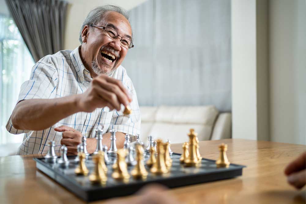 A smiling elderly man with glasses plays chess, holding a chess piece on a board with gold and silver pieces, sitting in a well-lit room that reflects the comfort of in-home senior care.