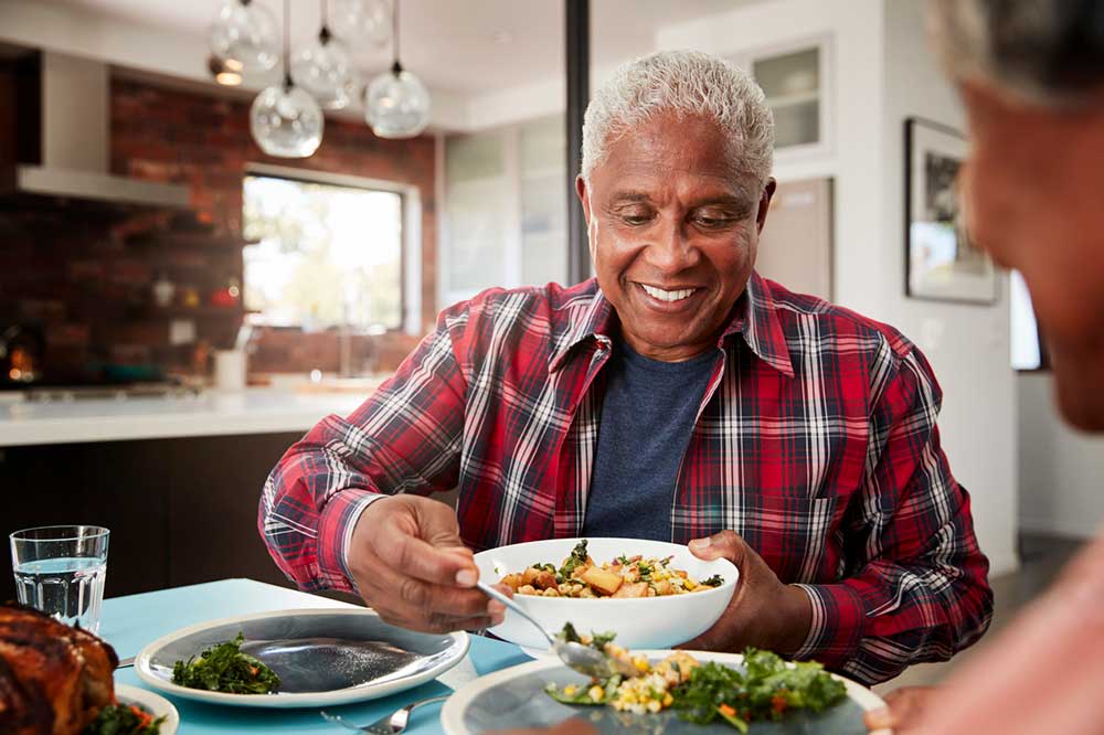 An elderly man wearing a plaid shirt smiles while serving food from a bowl at a dining table in a modern kitchen at United Church Homes.