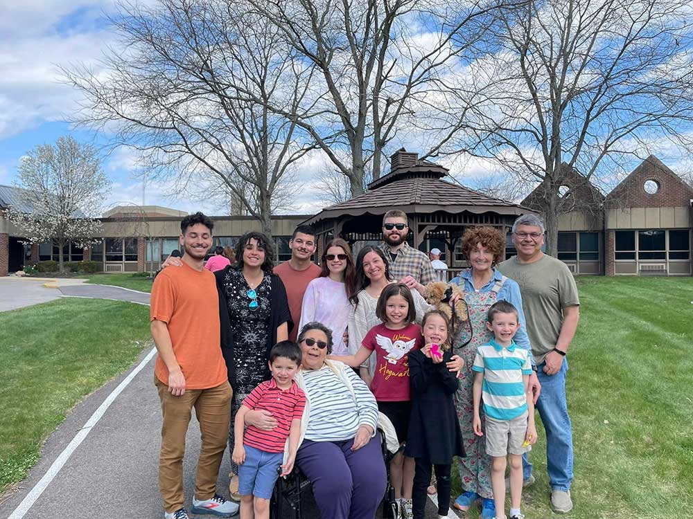 A group of people of varying ages stand together outdoors in front of a gazebo on a lawn, posing for a photo on a cloudy day, after giving reports at the community gathering.