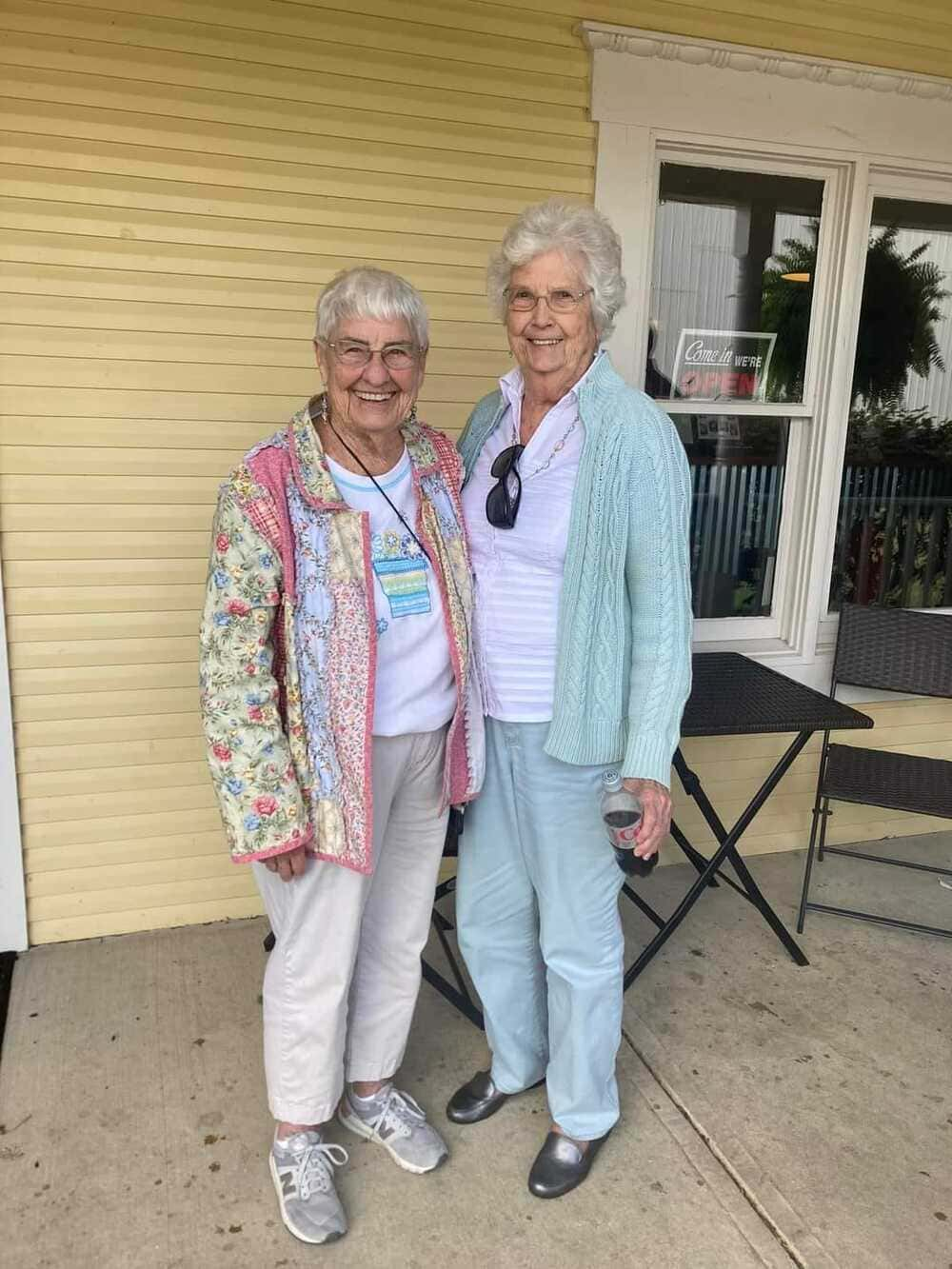 Two elderly women are standing in front of a yellow building, embodying the joy found in senior living options in Ohio. They both wear glasses and casual clothing, smiling warmly at the camera. A small table and chairs are visible in the background, adding to the cozy atmosphere.