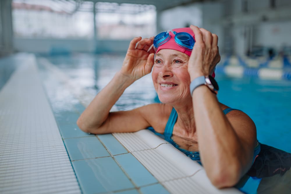 An elderly person, living in one of the vibrant independent living communities, is wearing a pink swim cap and blue goggles, smiling while holding the pool edge in an indoor swimming facility.