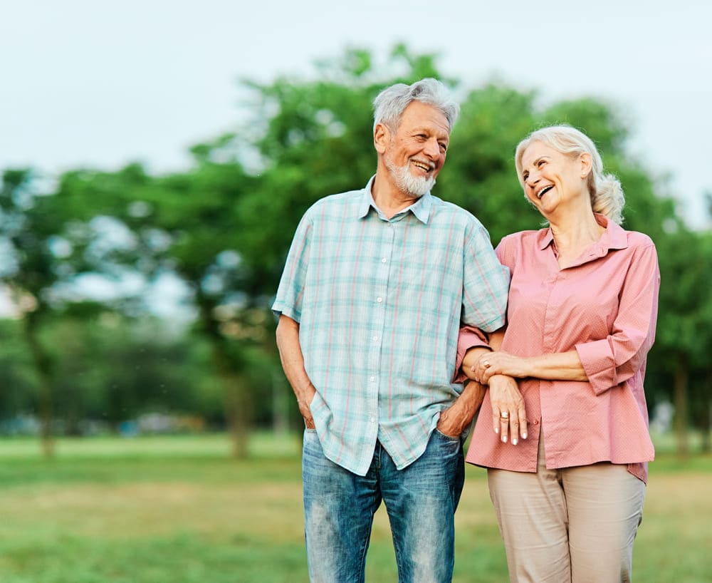 An elderly couple, a man in a plaid shirt and jeans and a woman in a pink blouse and pants, are walking arm-in-arm and smiling at each other in a park with green trees in the background, enjoying their time under the care of NaviGuide from United Church Homes.