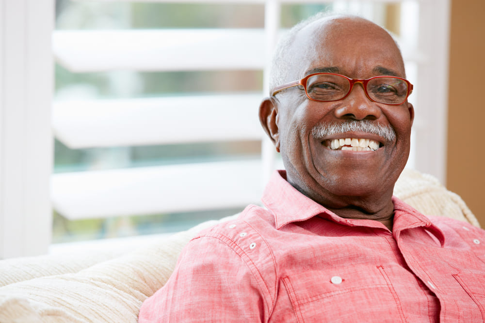 An elderly man with glasses and a mustache smiles while sitting on a couch, wearing a pink shirt. Blinds and windows are visible in the background, hinting at the cozy atmosphere of his skilled nursing facility in Ohio.