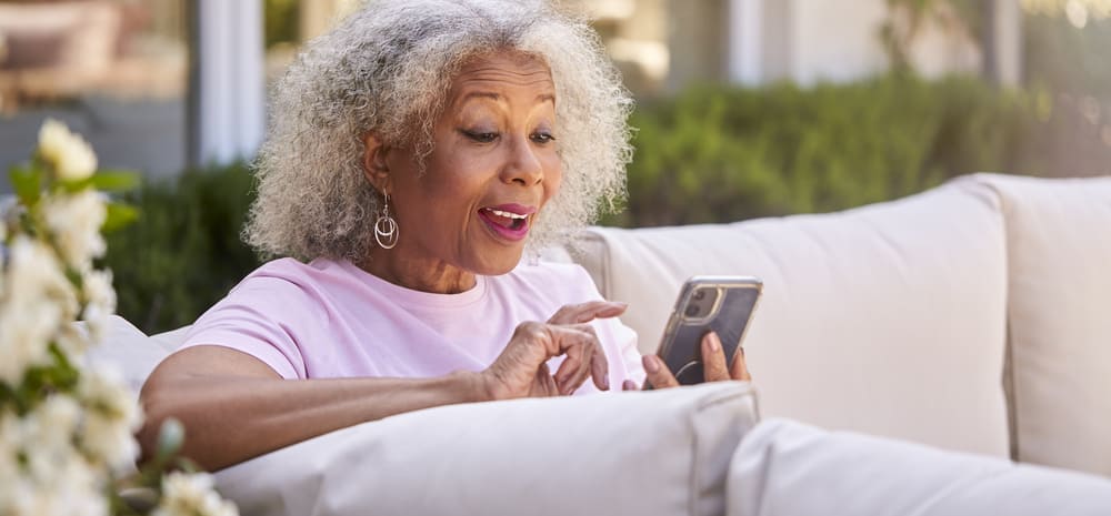 An elderly woman with curly gray hair is sitting on an outdoor sofa, looking at her smartphone and touching the screen with an expression of pleasant surprise as she reads the "About Us" section.