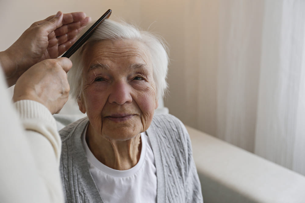 An elderly woman with short white hair is getting her hair brushed by someone else in a serene setting, emblematic of the compassionate hospice care in Ohio. She is sitting indoors and looking forward with a slight smile.