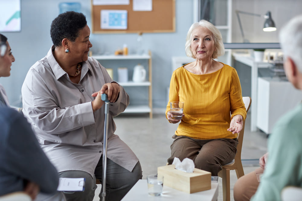 A group of people engaged in conversation sits in a circle; one elderly woman with a glass of water speaks while another woman with a cane listens attentively.