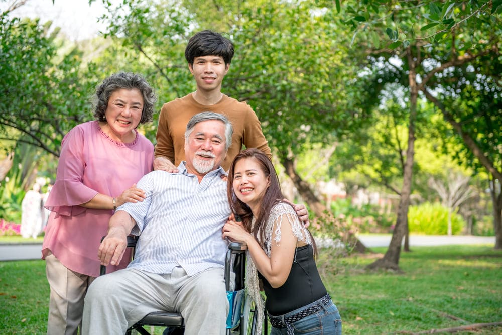 A smiling family poses outdoors in a park. An elderly man, actively participating in stroke rehab, is surrounded by three people: two women and one young man. Trees and greenery can be seen in the background.