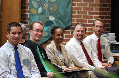 Five people are sitting in a row, dressed in business attire, with name tags. They are against a brick wall with a green, leafy decorative textile hanging.