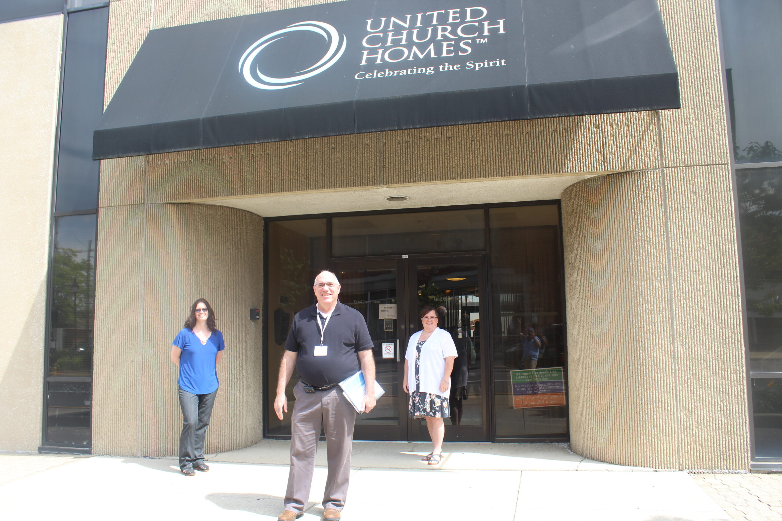 Three people stand outside the entrance of a building with a sign that reads "United Church Homes: Celebrating the Spirit." The awning is black, and the building has tan walls with large windows. Among them is the director of procurement and supply chain, engaging in warm conversation.