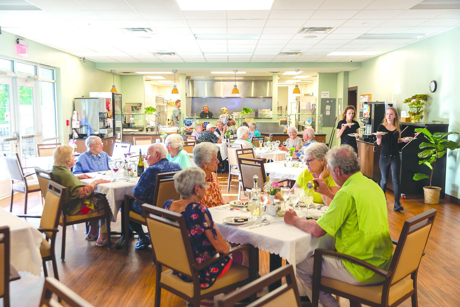 A group of elderly individuals dining in a well-lit cafeteria, with several tables set for the meal, while staff members stand near a podium at the front of the room, fostering a culture change towards community and engagement.