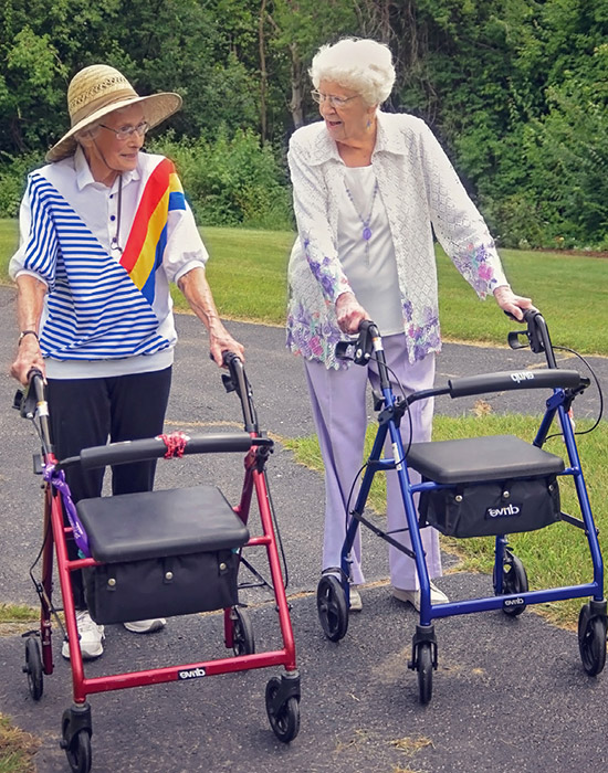 Two elderly women are walking side by side on a paved path, each using a rollator walker. They are outdoors, surrounded by greenery, and appear to be conversing.