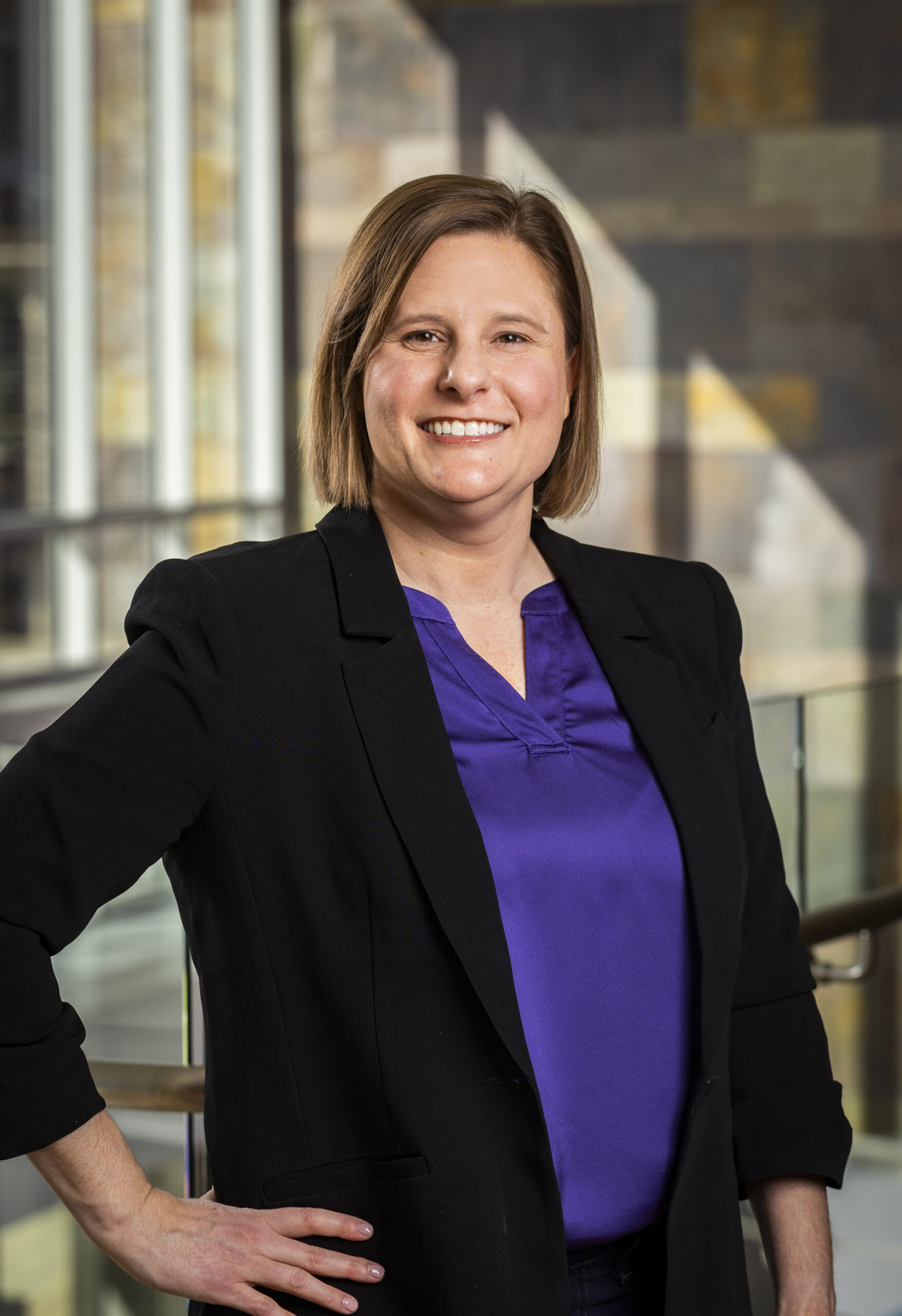 A person stands indoors wearing a black blazer and purple shirt, exuding leadership while smiling at the camera with hands on hips.