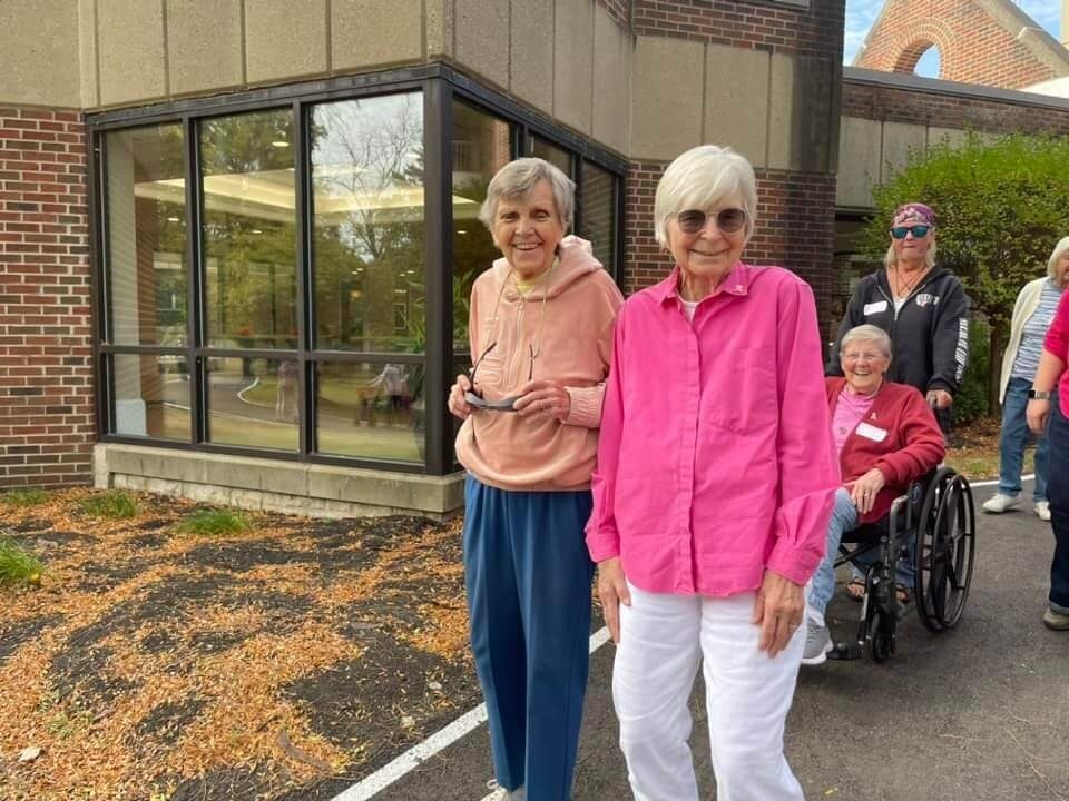 A group of elderly individuals, two standing and two being pushed in wheelchairs, are outside a brick building on a sunny day. The two standing women from the assisted living community are smiling at the camera.