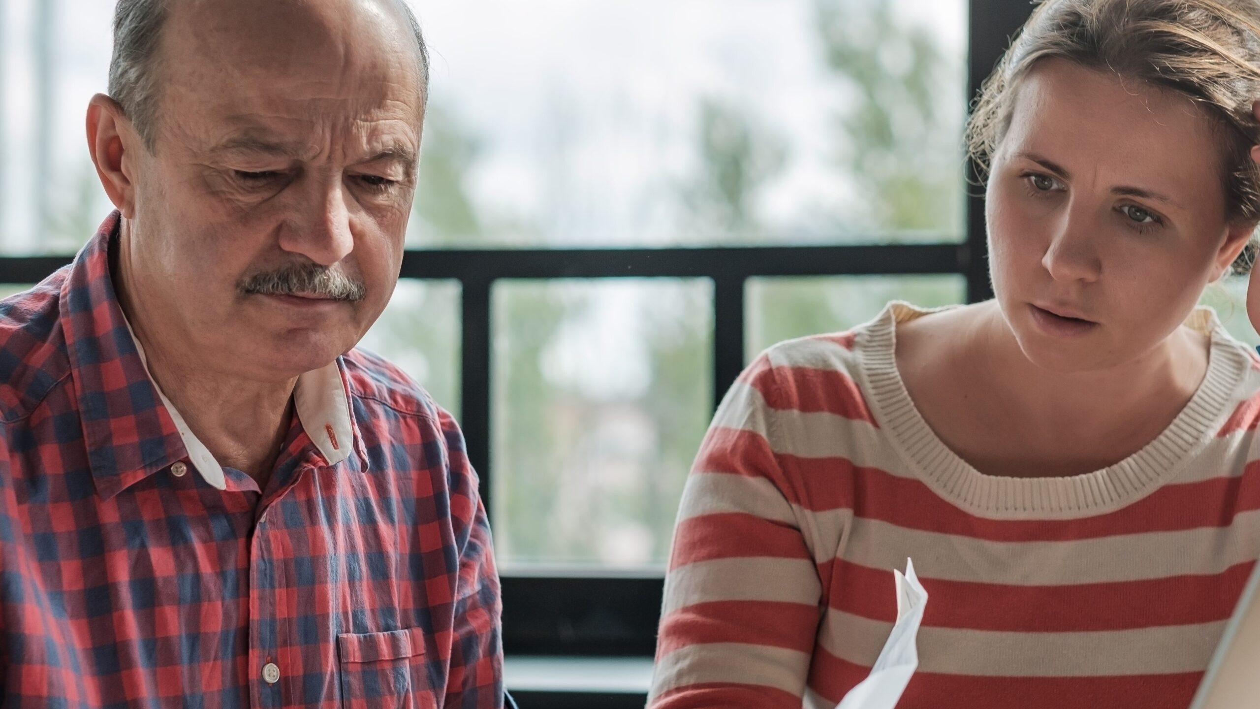 An older man in a plaid shirt and a younger woman in a striped sweater are reviewing documents together by a window.