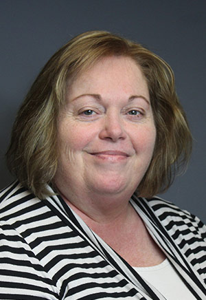 A woman with short, light brown hair wearing a black-and-white striped top and a white shirt, part of our dedicated fundraising team, smiles at the camera against a gray background.