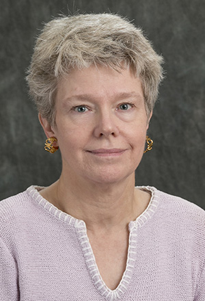 A person with short gray hair, wearing a light pink sweater and gold earrings, poses against a dark blurred background, reminiscent of a dedicated member of a fundraising team.