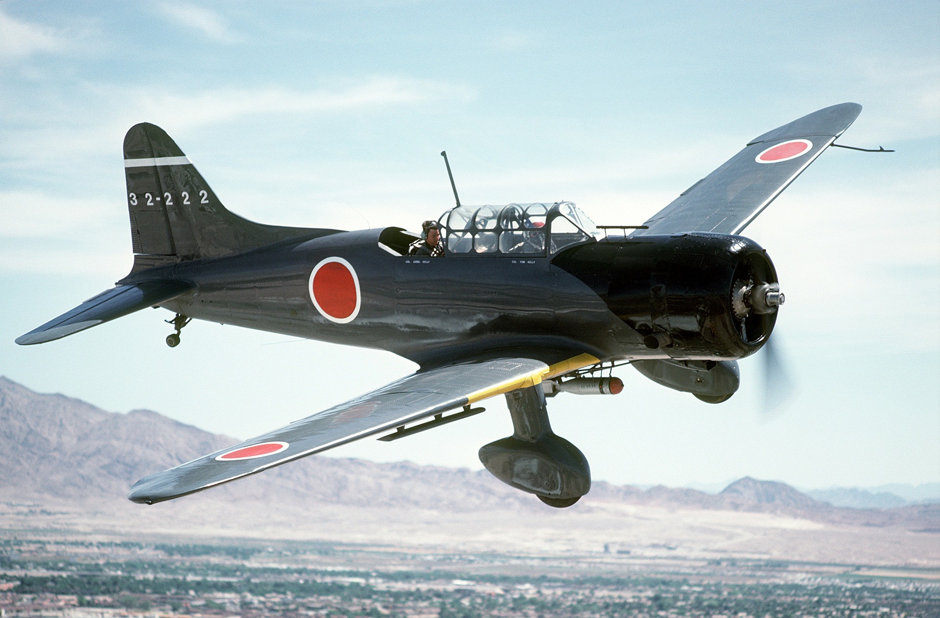 A black World War II-era Japanese fighter plane with red and white markings is flying over a mountainous landscape.