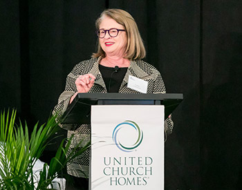 A woman is speaking at a podium with a sign that reads "United Church Homes." She is wearing glasses and a patterned jacket.