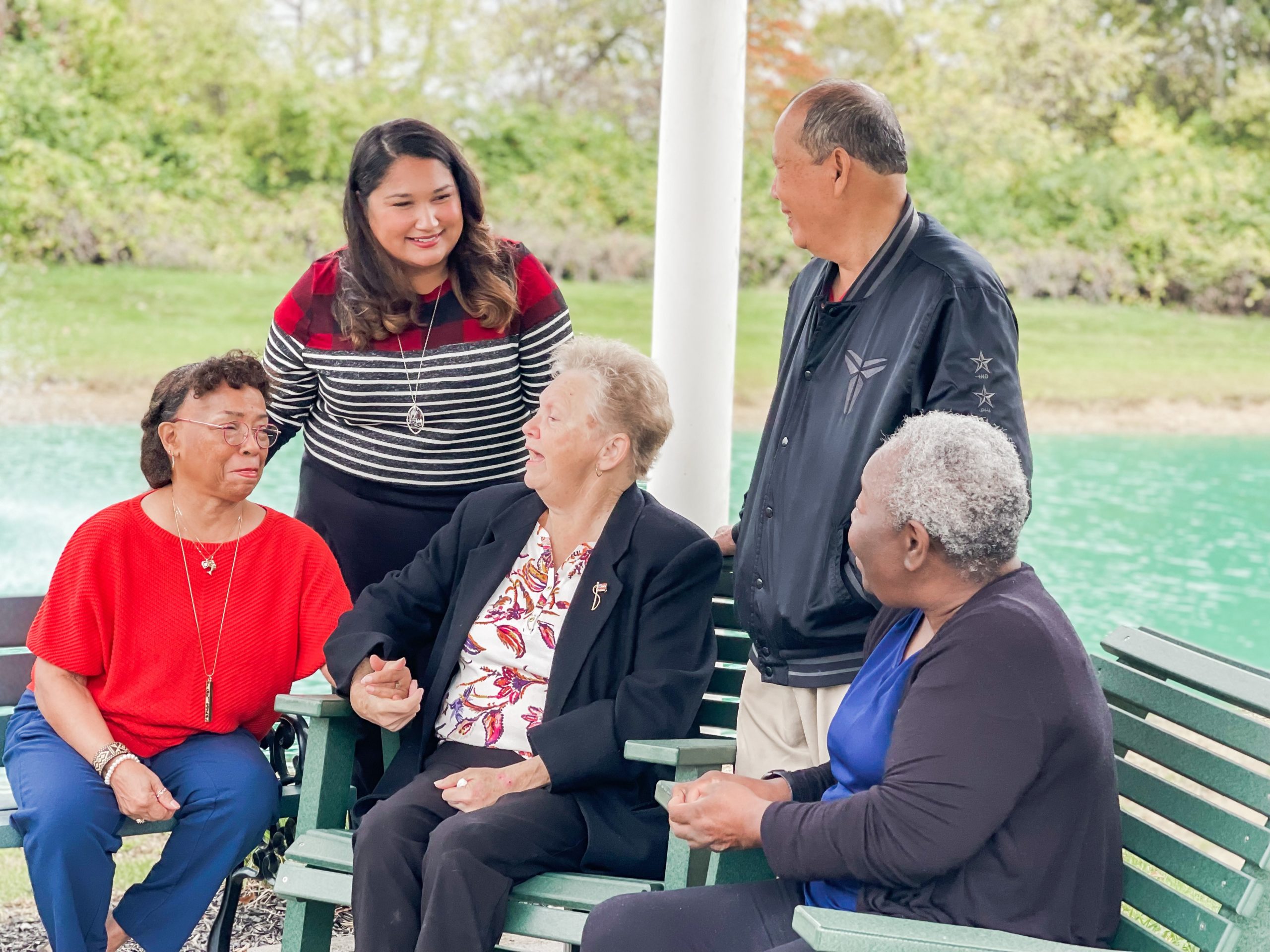 A group of five people, four seated on a bench and one standing, engage in conversation near a pond in an outdoor setting.