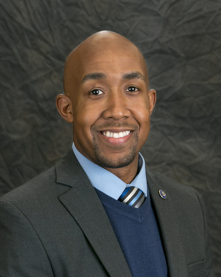 A smiling man in a suit and tie poses for a professional headshot against a dark, crumpled background, exuding leadership and confidence.