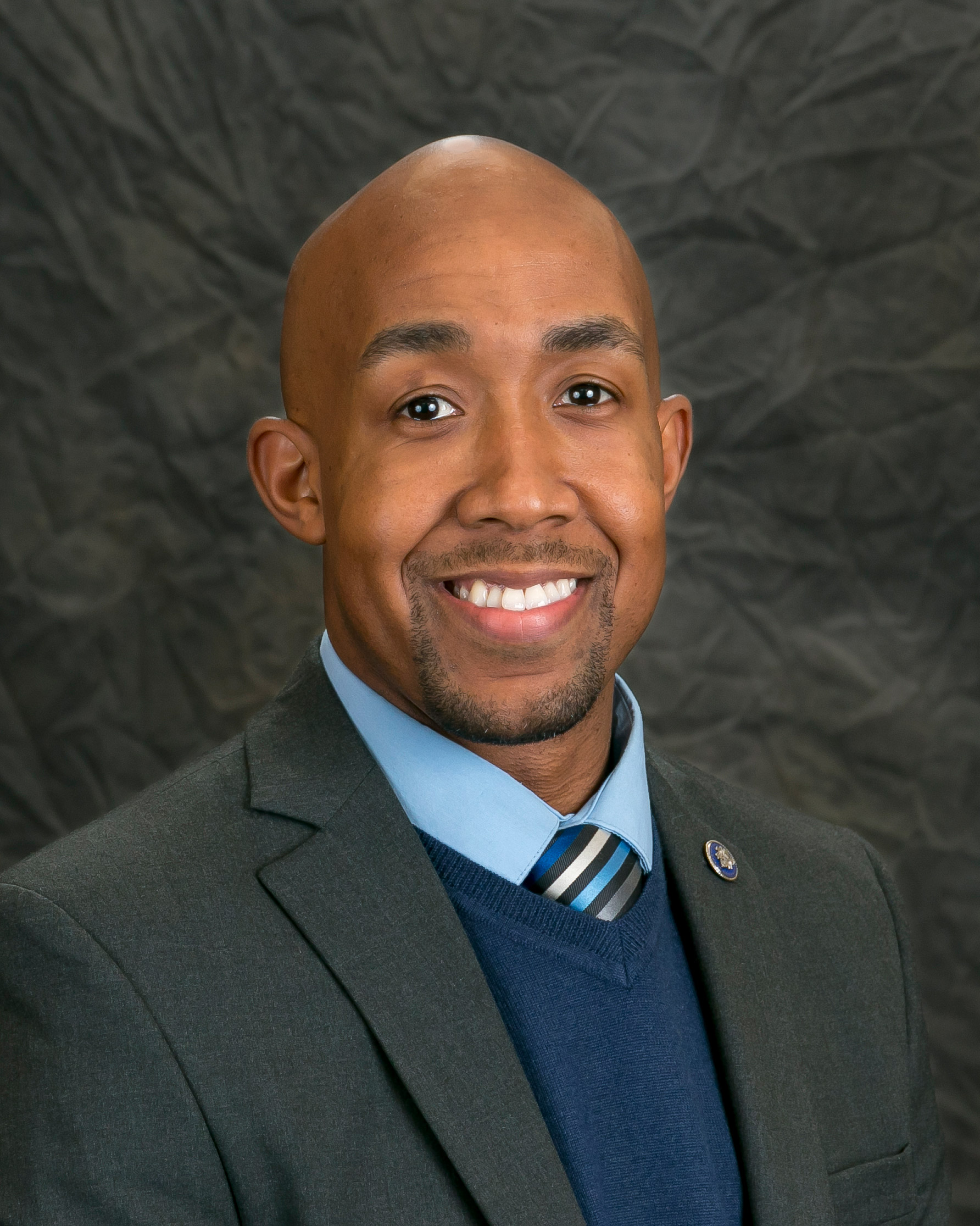 A bald man in a suit jacket, blue shirt, and striped tie smiles at the camera against a textured dark background.