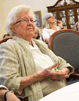 An elderly woman with gray hair, wearing glasses and a light-colored outfit, sits in a room with other people, looking forward with a neutral expression.