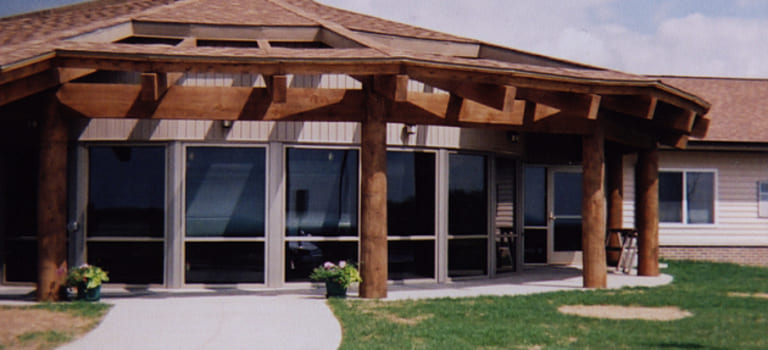 A building with large windows and wooden columns supports a sloped roof. The scene includes a concrete walkway and grass lawn under a partly cloudy sky, representing affordable housing in Ogema, Minnesota.