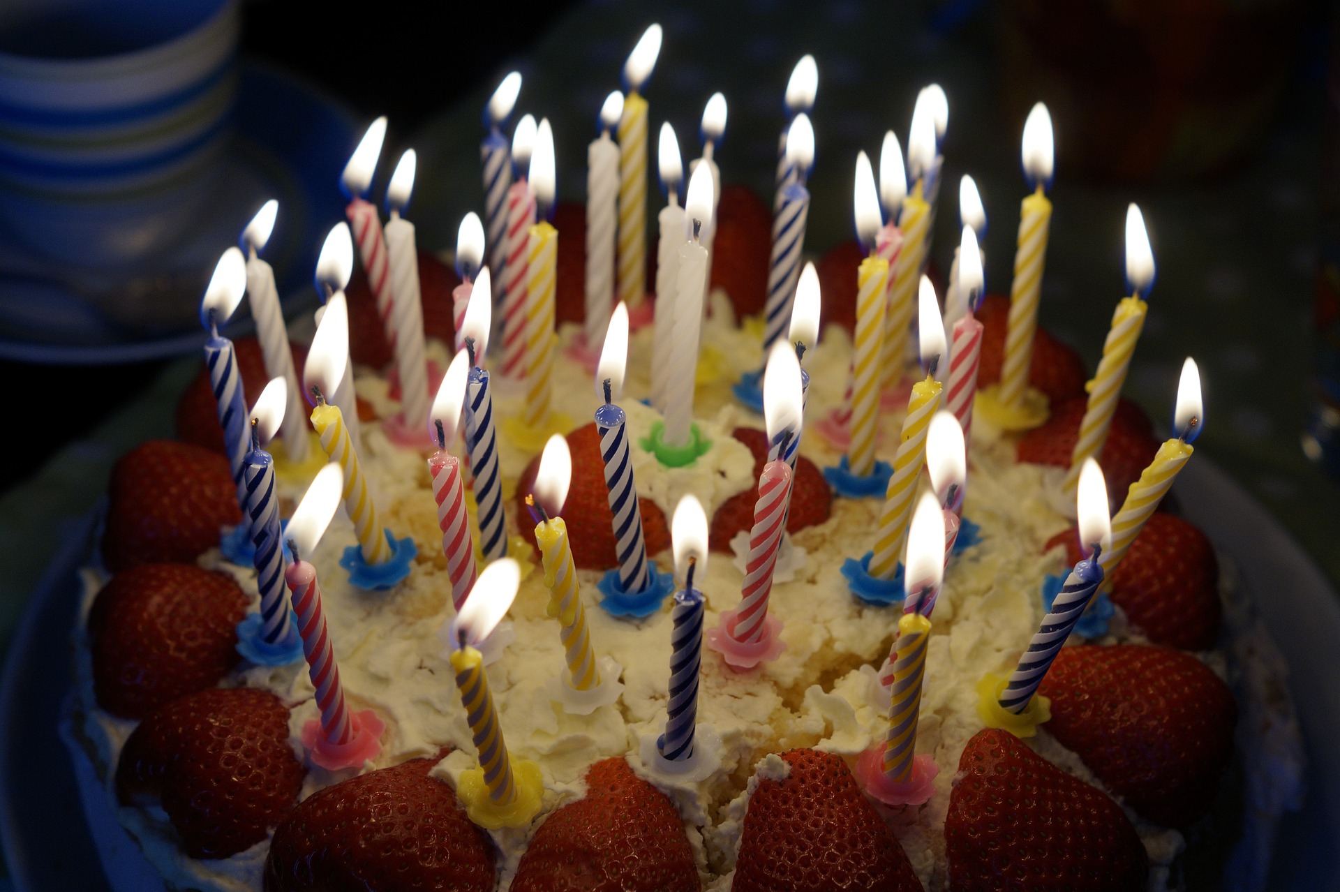 A birthday cake topped with lit candles and strawberries. The cake is decorated with a mix of yellow, blue, pink, and striped candles. Plates and cups are visible in the background.