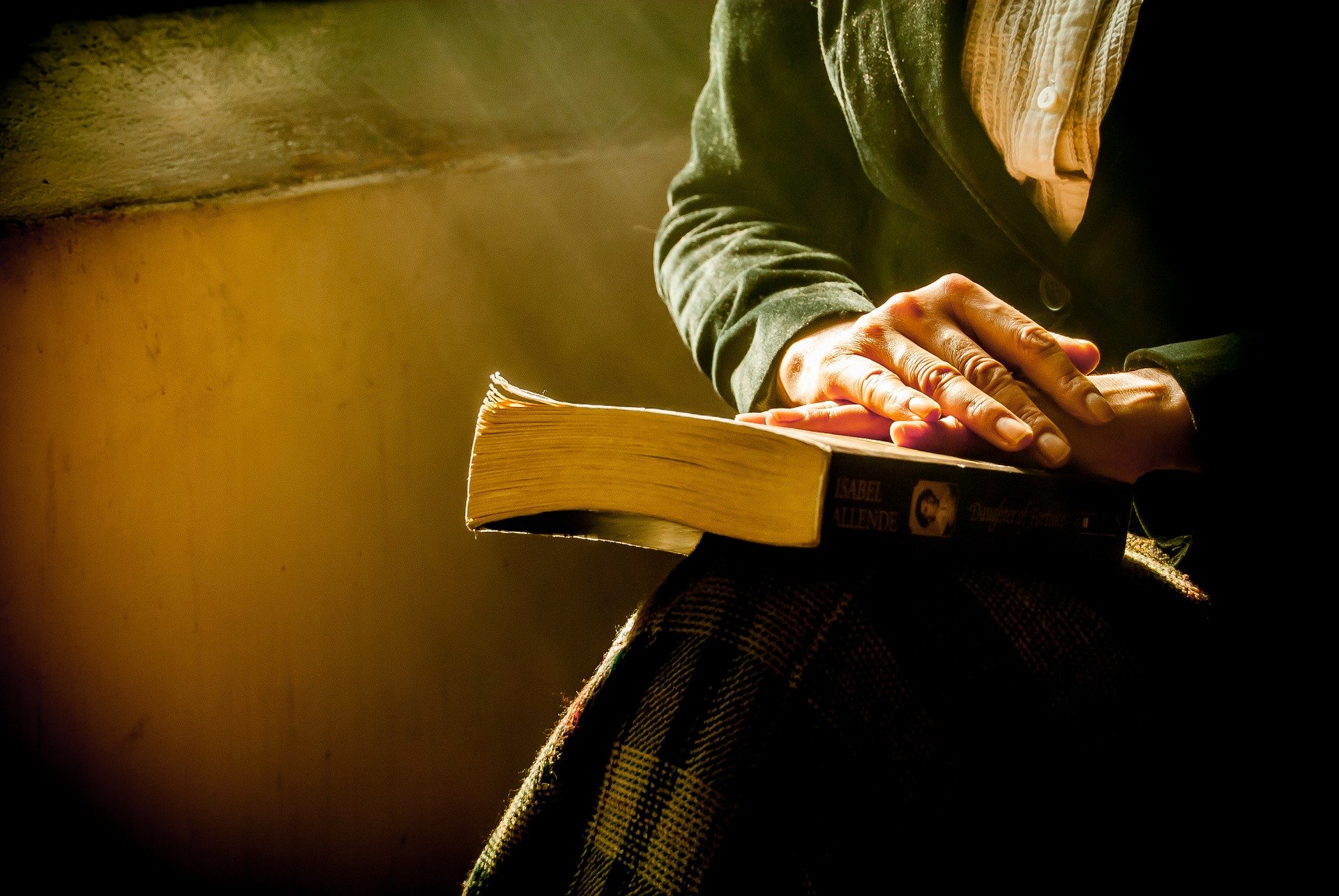 A person holding a book while sitting by a window with sunlight streaming in. Their hands rest on the book's cover, and they are wearing a plaid skirt and a dark jacket.