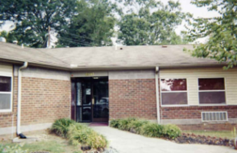 A small brick building in Oxford with a single entrance, flanked by two windows on each side. A narrow sidewalk leads to the entrance, embodying the charm of Mississippi. Trees and shrubs are present in the background, contributing to an inviting atmosphere for affordable housing in Oxford.