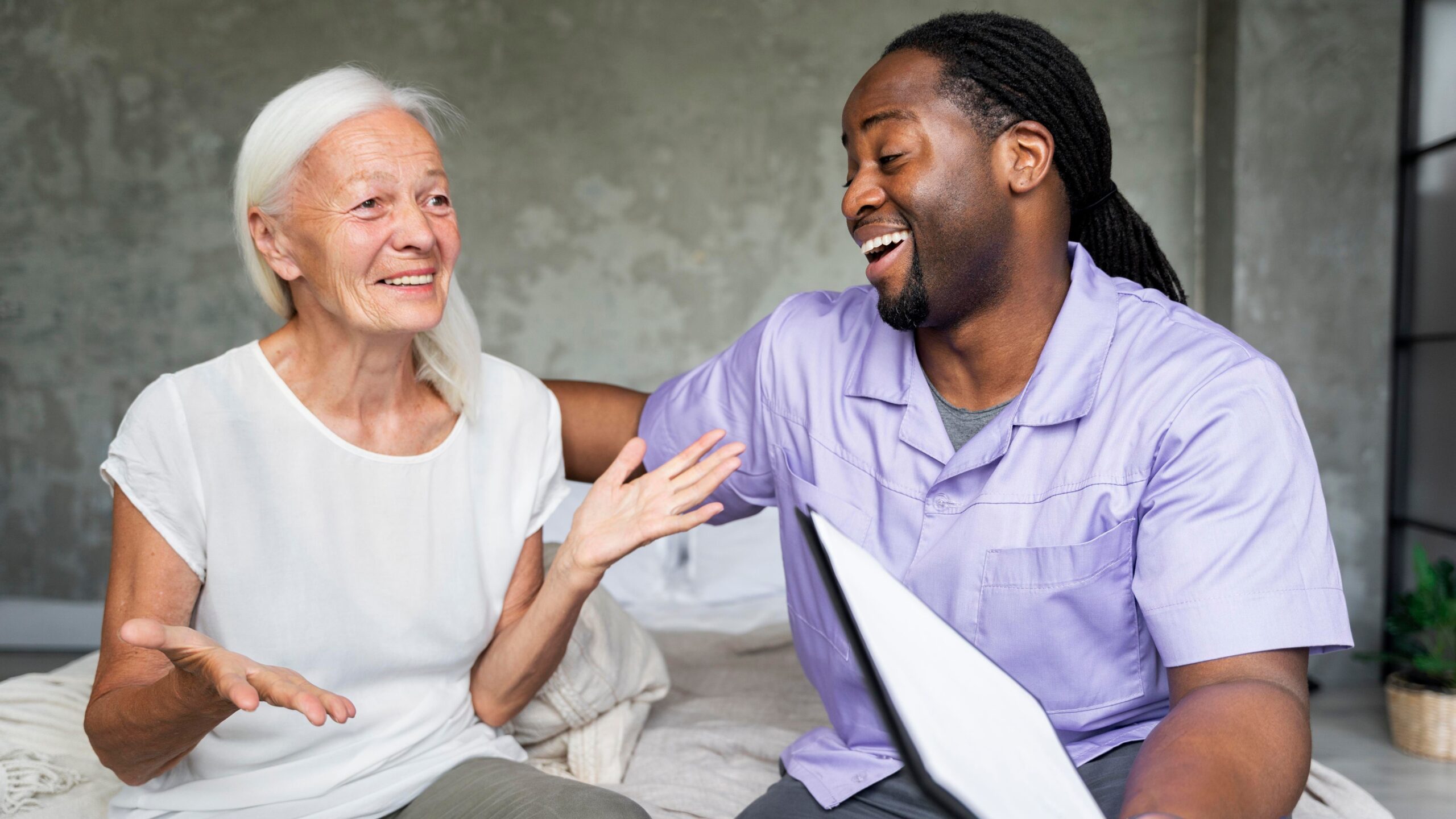 An elderly woman and a caregiver share a light-hearted moment while engaged in conversation, both smiling and gesturing expressively.