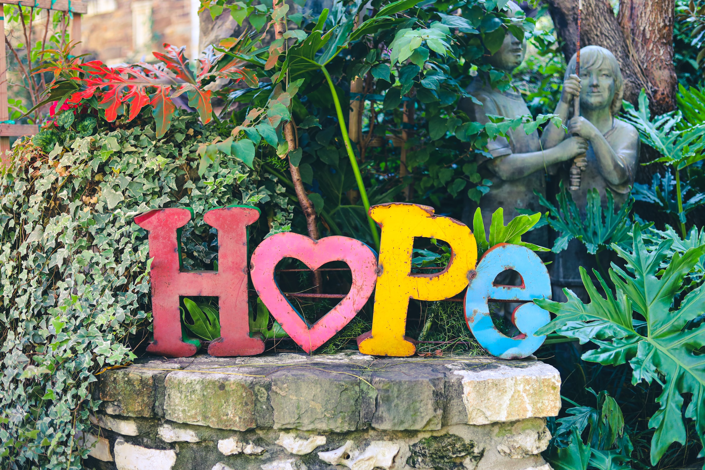 Colorful letters spelling "Hope," with the 'O' shaped as a heart, are placed on a stone ledge surrounded by lush green foliage and garden statues.