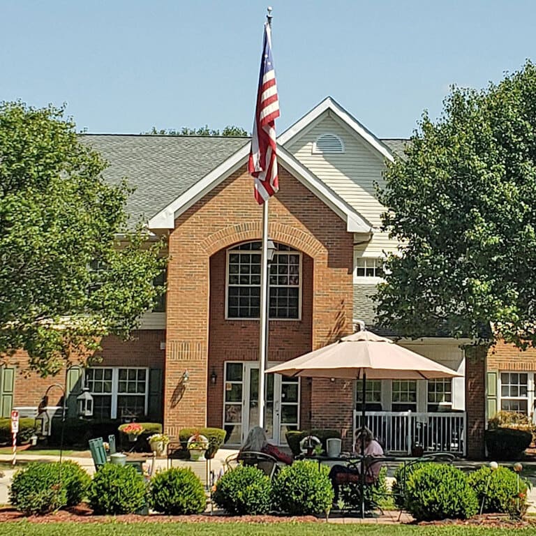 A two-story brick building with a large American flag in front stands as an example of affordable housing in North Lewisburg, Ohio. A patio area adorned with tables, chairs, and an umbrella is situated near the entrance, surrounded by lush greenery and trees.