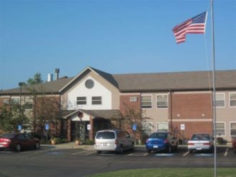 Facade of a two-story building with a mix of brick and siding, featuring a parking lot with several cars in front and an American flag flying on a pole. This charming example of affordable housing is located in Fredonia, New York.