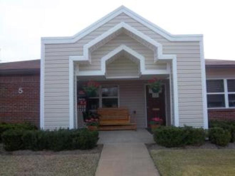 A small, single-story brick and siding building with a covered entrance, wooden bench, potted flowers, and manicured shrubs. The facade of this affordable housing in Greenwood, Mississippi features an intricate geometric design above the entryway.