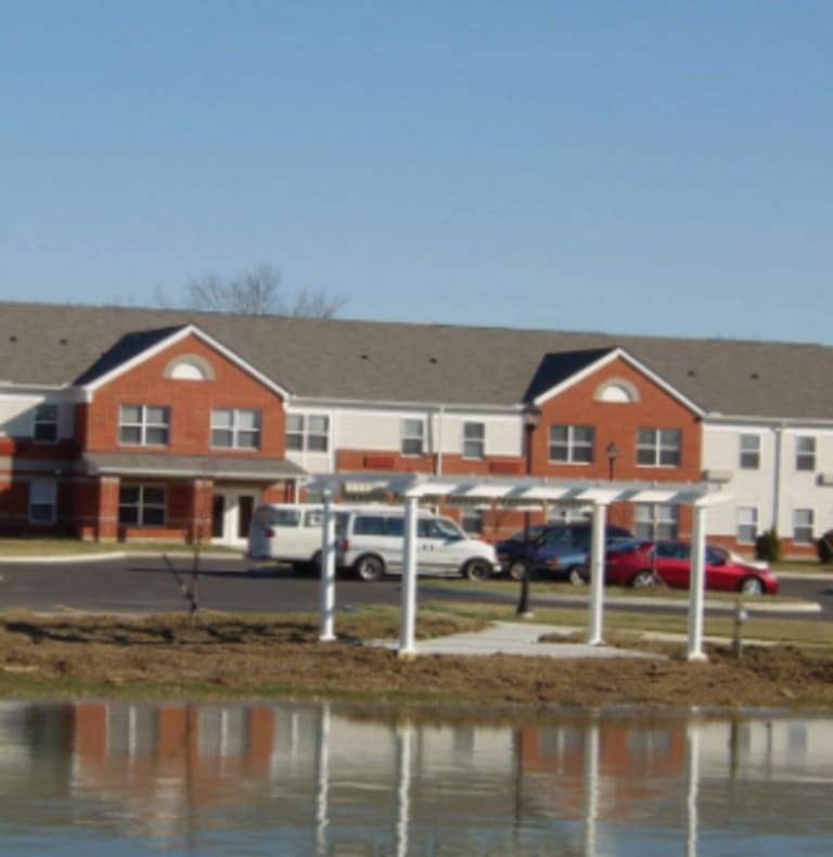 Two-story brick and siding buildings with white trim near a small pond offer affordable housing in Canal Winchester, Ohio. Several cars are parked by the buildings, and a white pergola is situated near the water under a clear sky.