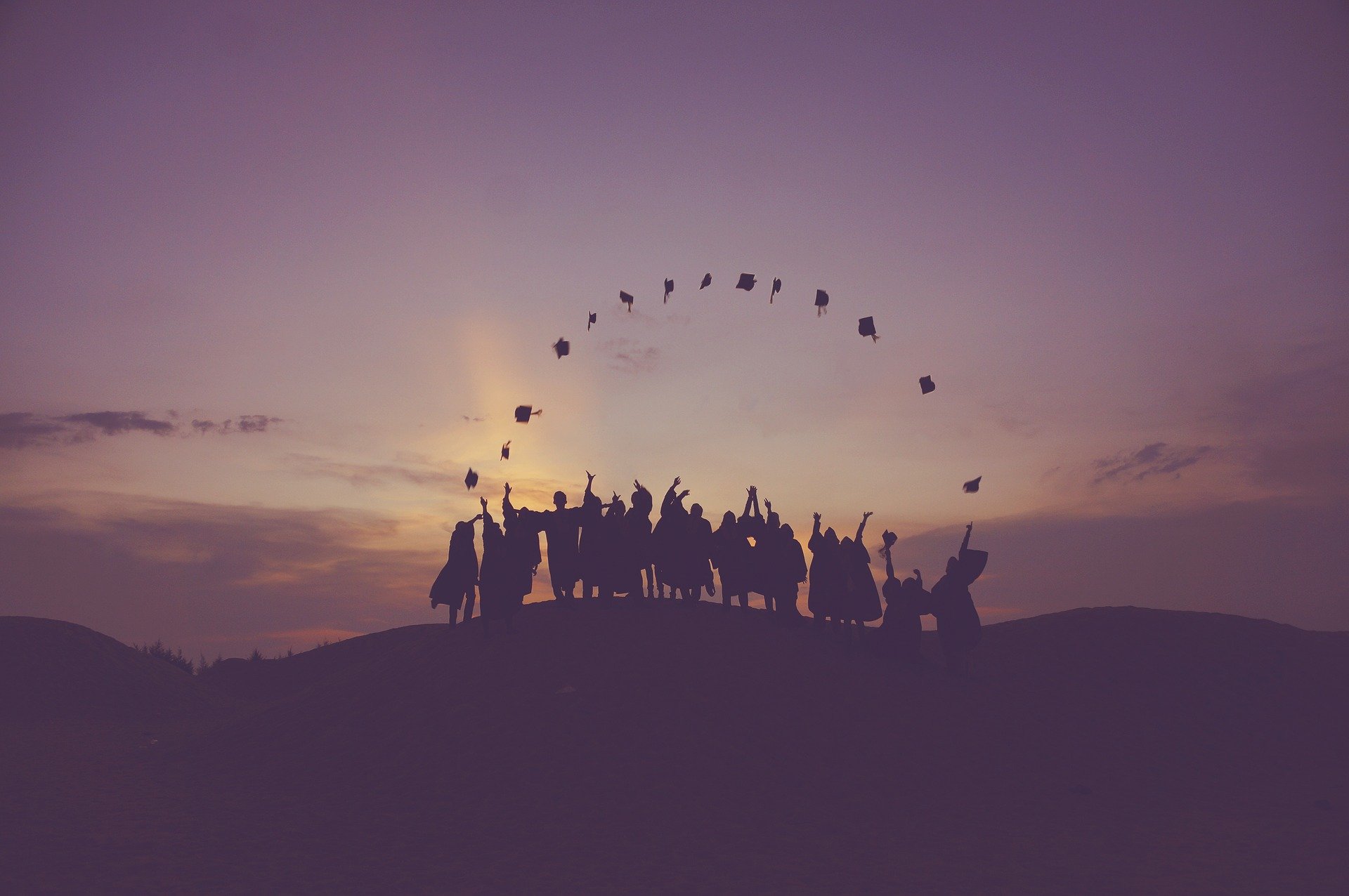 A group of people silhouetted against a sunset sky throws graduation caps into the air while standing on a hill.