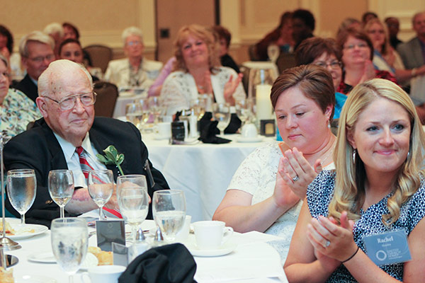 An elderly man in a suit sits at a banquet table, surrounded by people listening attentively and clapping. Tables are set with glasses, cups, and plates. A woman with a nametag reading "Rachel" is in the foreground.