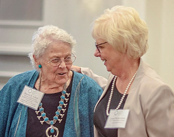 Two older women, both wearing glasses and name tags, stand close together, smiling. One wears a blue outfit with a large turquoise necklace, and the other wears a beige jacket and pearl necklace.