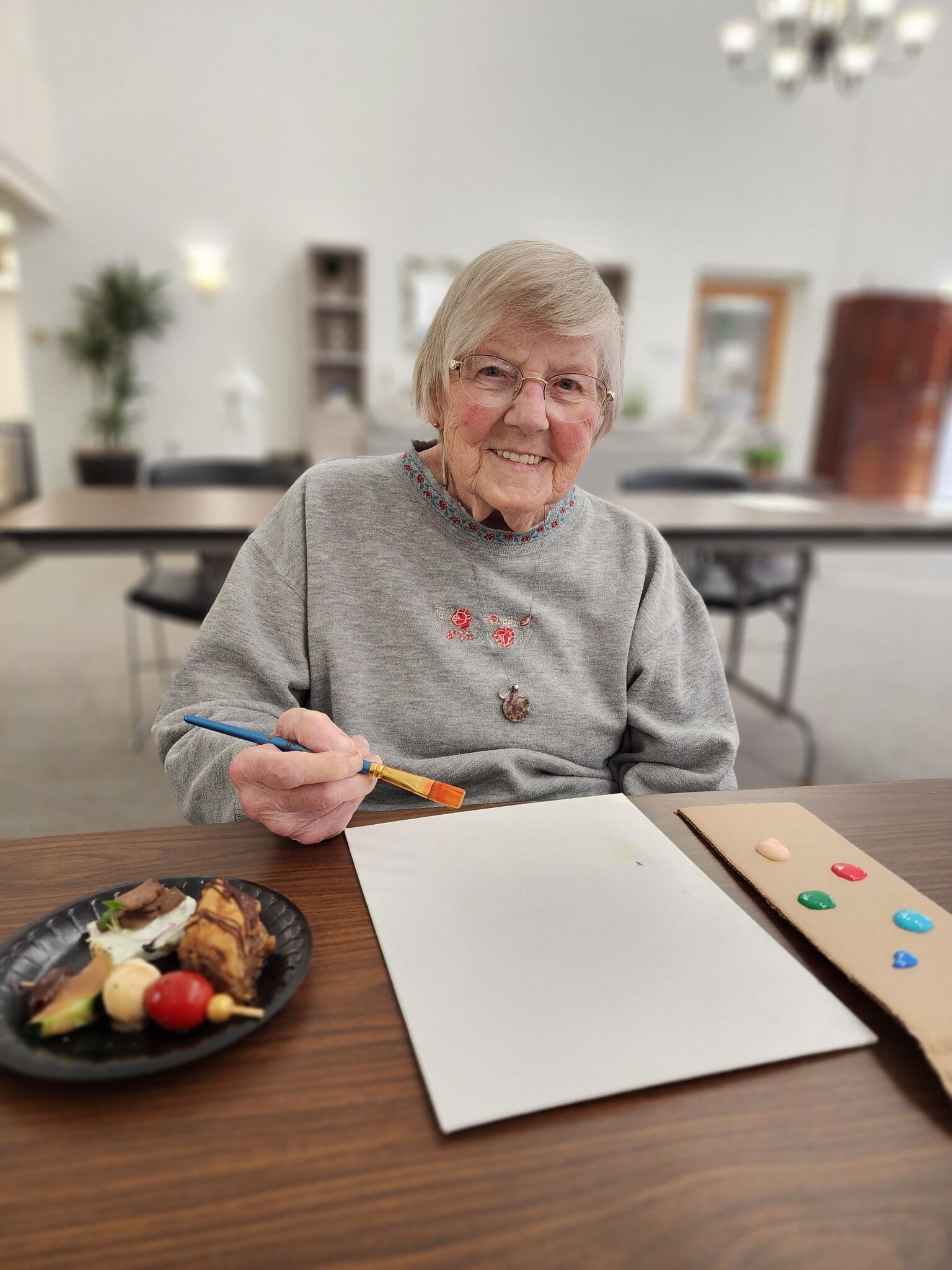 Elderly woman seated at a table, holding a paintbrush over a blank sheet of paper, with a plate of food to her left, embodying the vibrant life celebrated by careers at United Church Homes.