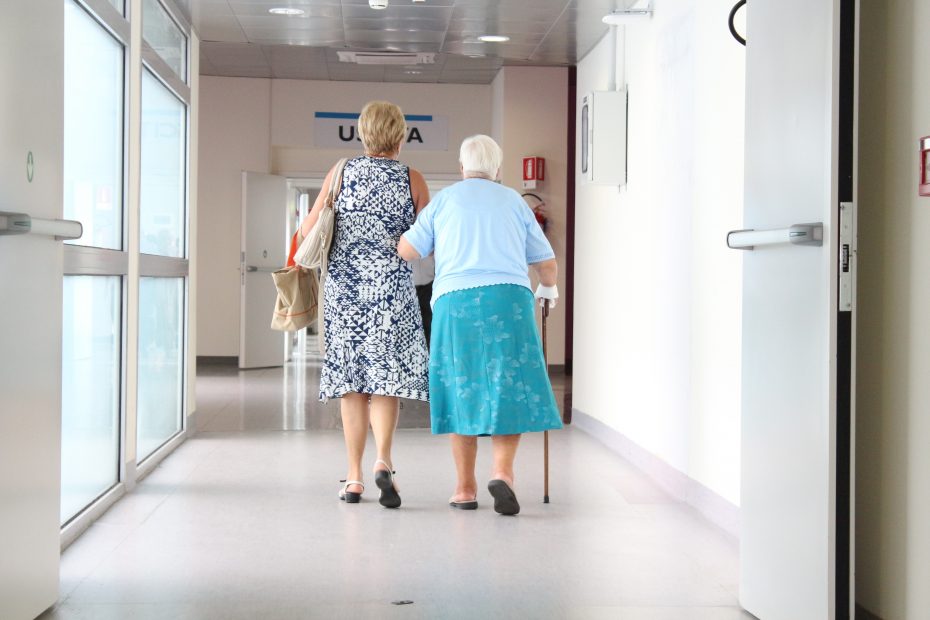 Two elderly women walk down a hospital corridor; one uses a cane while the other supports her arm.